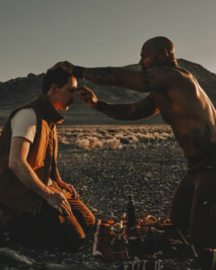 Man in a vest sitting on the desert ground while another man places a hand on his head