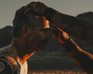 Close-up profile of a man with a tattooed arm as another hand rests on his forehead