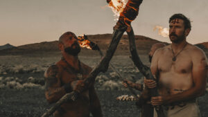 Two men holding crossed flaming torches in the desert at sunset