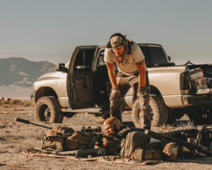 Man in ear protection standing beside a pickup truck in the desert with gear laid out on the ground