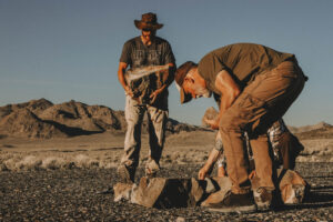 Two men in the desert, one standing in a hat and one bent over working on the ground
